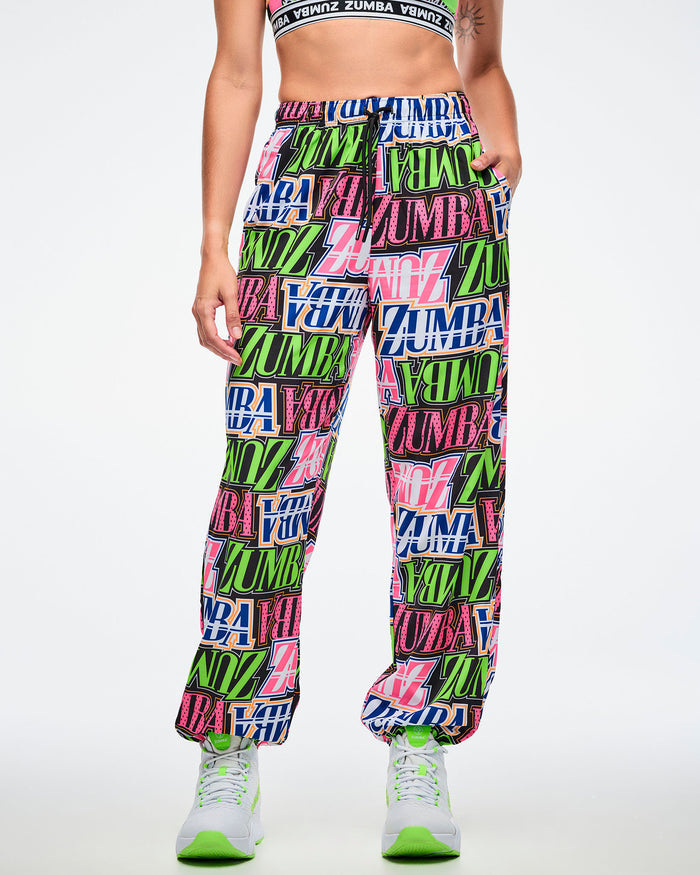 Front view of a female model with her hand in her left pocket, wearing colorful Zumba pants paired with green and white shoes on a white background.