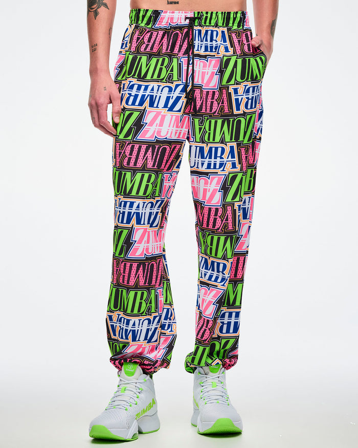 Male model with his left hand in his pocket, wearing multicolored woven pants paired with white and green shoes.