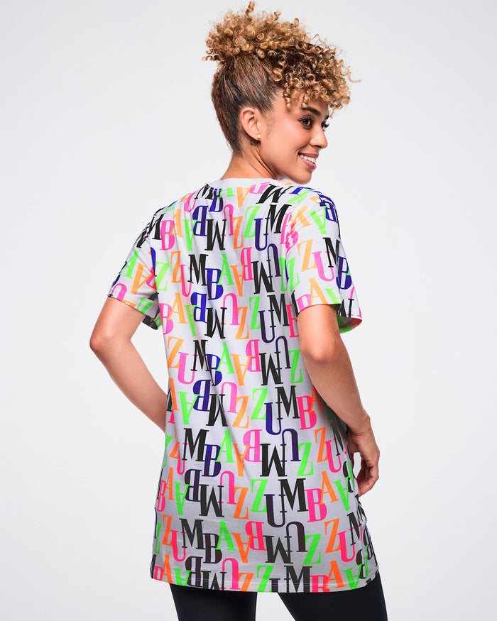 Back view of a female model wearing a colorful 'ZUMBA' patterned t-shirt against a white background.