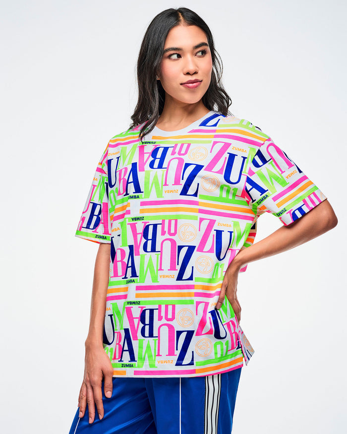 Female model wearing a colorful Zumba T-shirt with one hand at her side, paired with blue Zumba pants.