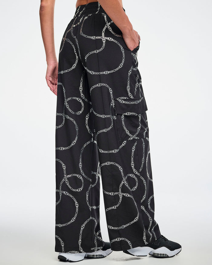 Back view of the black cargo pants with silver chain pattern worn by a female model and paired with black and white shoes.
