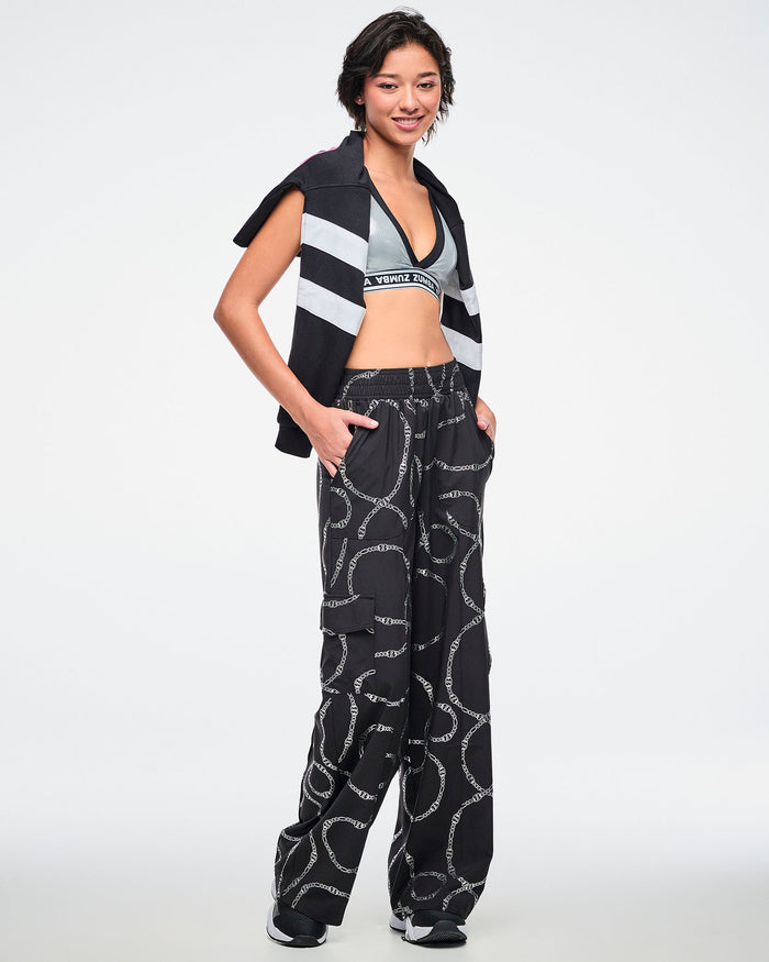 Female model wearing black and silver chain-patterned cargo pants, paired with a silver bra and a black-and-white polo draped over her shoulders.