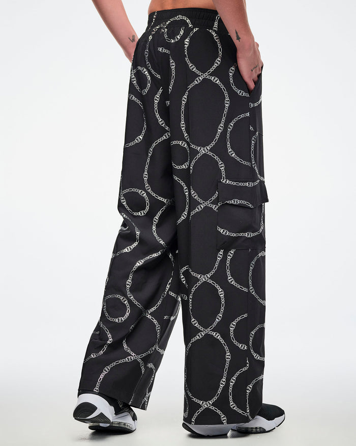 Back view of a male model wearing black cargo pants with silver chain pattern, paired with black and white shoes.