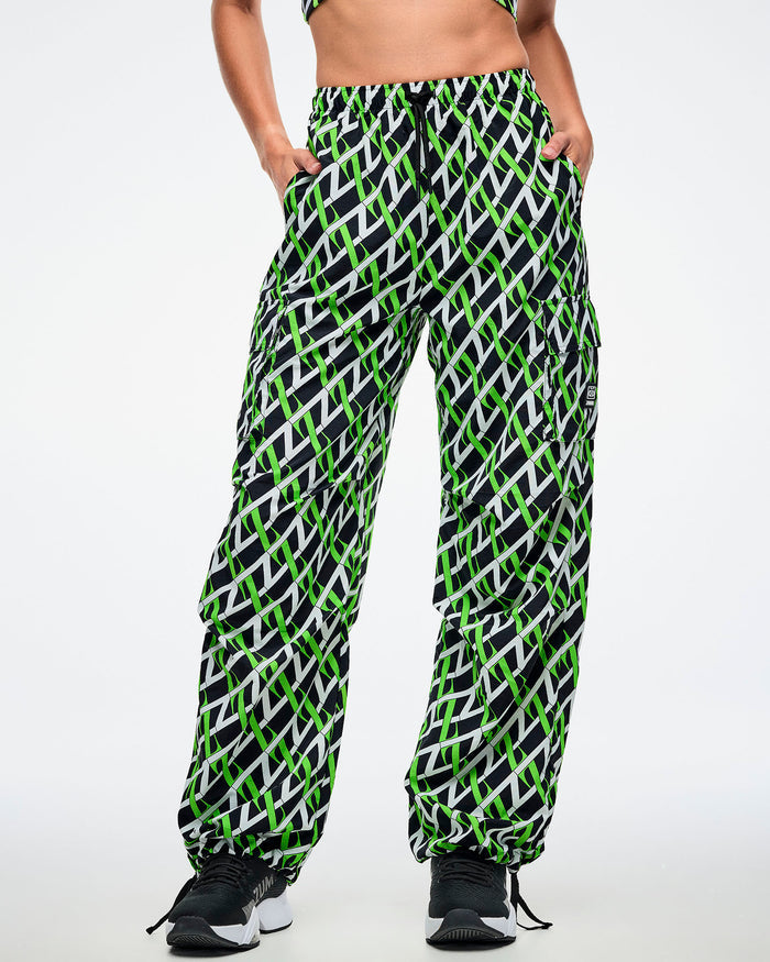 Front view of a female model wearing cargo pants with an allover black, white and neon green “Z” crisscross pattern, left hand in her pocket, paired with black-and-white shoes.