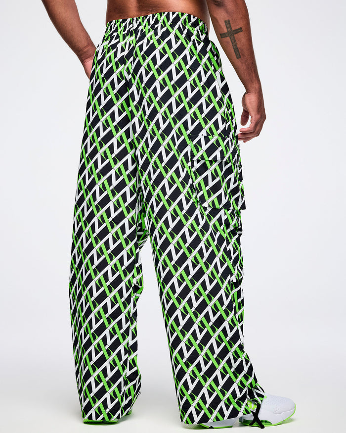 Back view of a male model wearing black cargo pants with an allover black, white and green “Z” crisscross pattern, left hand in his pocket, paired with black-and-white shoes.