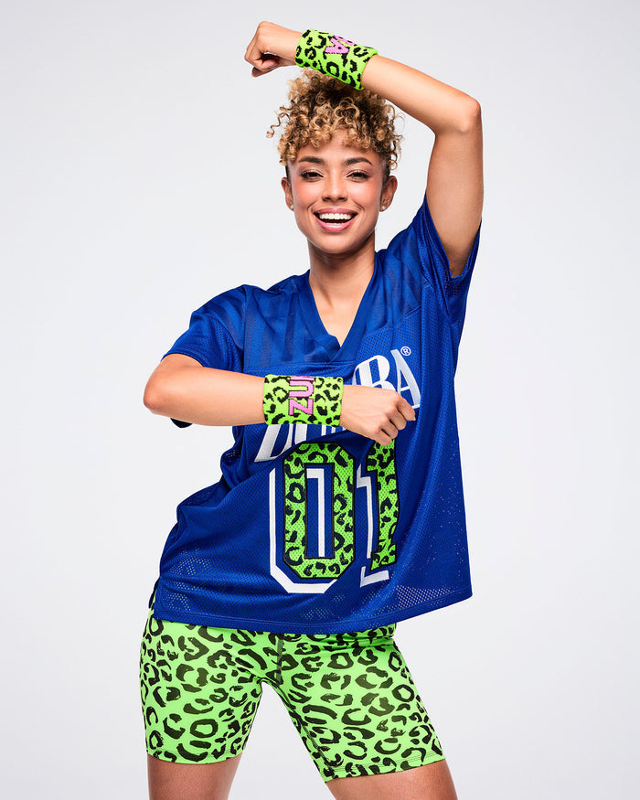 Female model wearing matching green-and-black leopard-print wristbands with “Zumba” in pink lettering, paired with a blue top and green-and-black leopard-print biker shorts.