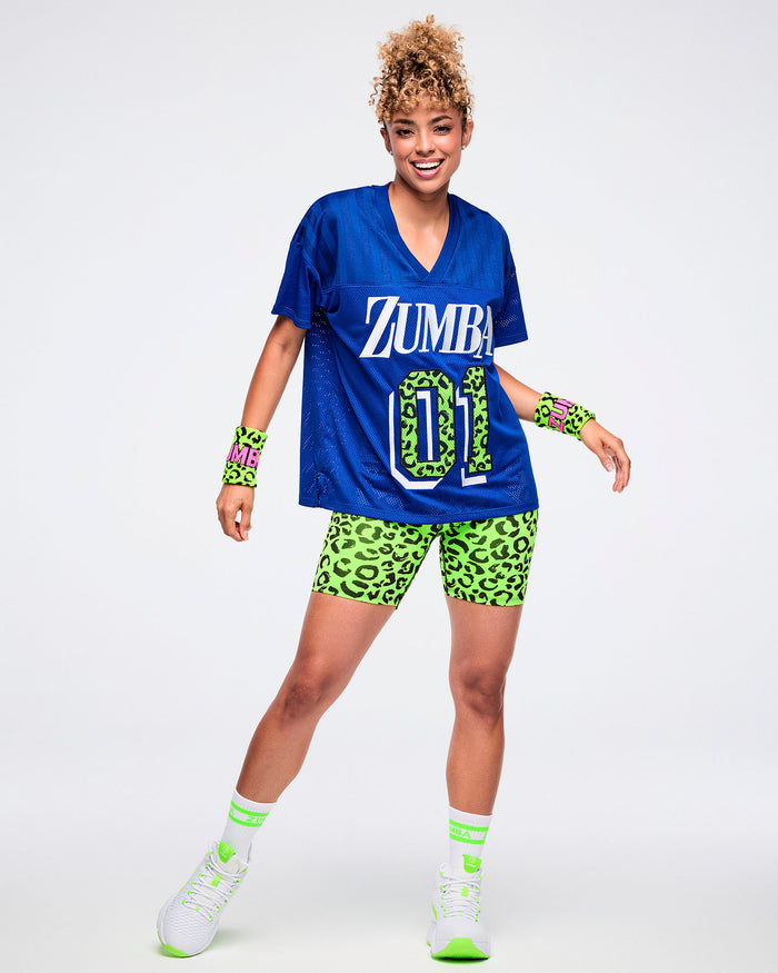 Full view of a female model wearing matching green-and-black leopard-print wristbands with “Zumba” in pink lettering, paired with a blue top and green-and-black leopard-print biker shorts and green and white socks and shoes.