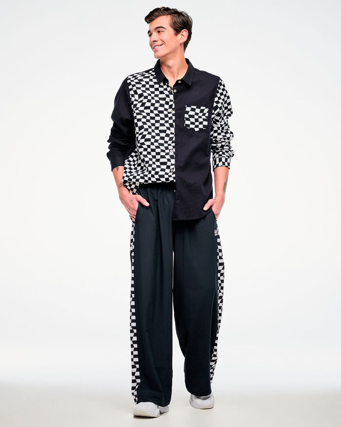 Full view of a male model wearing a black and white checkered flannel button-up, paired with black pants featuring a checkered side stripe and white shoes.
