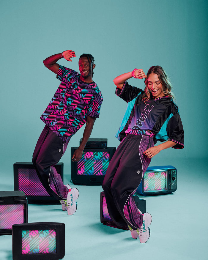 A male and female model dancing in matching streetwear outfits with retro TV props against a teal studio background. The outfits feature a black base with vibrant pink, purple, and aqua graphic patterns and "ZUMBA" text.