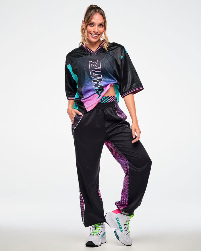 Female model wearing a black jersey tee with a pink-to-purple gradient "ZUMBA" print, paired with black joggers and white sneakers.