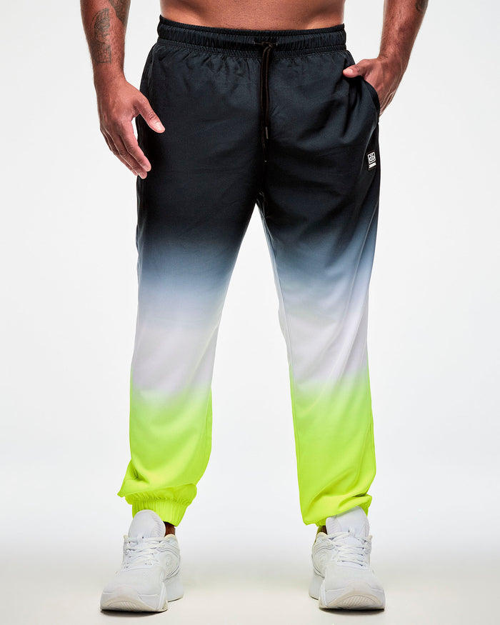 Male model wearing black, grey, white, yellow ombre joggers with a drawstring waistband and paired with white sneakers, hand in the side pocket.