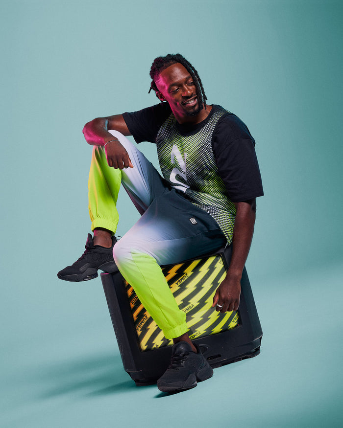 Male model wearing green and black dot "ZUMBA" tank and black-to-neon-yellow ombre joggers, paired with black shoes, sitting on an old 80's style television against a blue studio background.