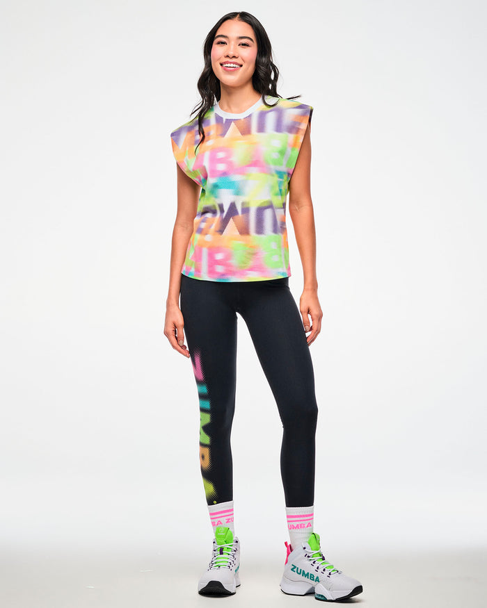 Full view of a female model wearing an multi-color extended shoulder tank with a blurred wordmark print, paired with black ZUMBA leggings, pink striped socks, and white sneakers.