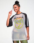 Female model wearing a white mesh oversized tee with black and white striped sleeves, a big yellow and orange Zumbito logo and "ZUMBA" text in black and orange above it, all paired with black and yellow dot-print leggings.
