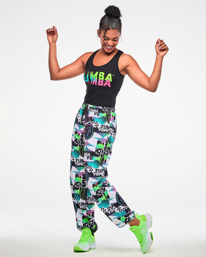 Female model wearing a black ribbed tank, paired with white multicolor "ZUMBA" print pants and green sneakers, dynamic dance pose.