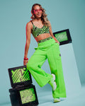 Female model wearing green track pants and a green and black geometric striped print bra, paired with white shoes against a light blue background standing next to retro televisions.