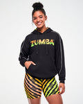 Female model wearing a black hoodie with an ombre orange, yellow, and green "ZUMBA" logo and thin white stripes down the sleeves, paired with matching lightning bolt-print biker shorts, hand in the hoodie kangaroo pocket.
