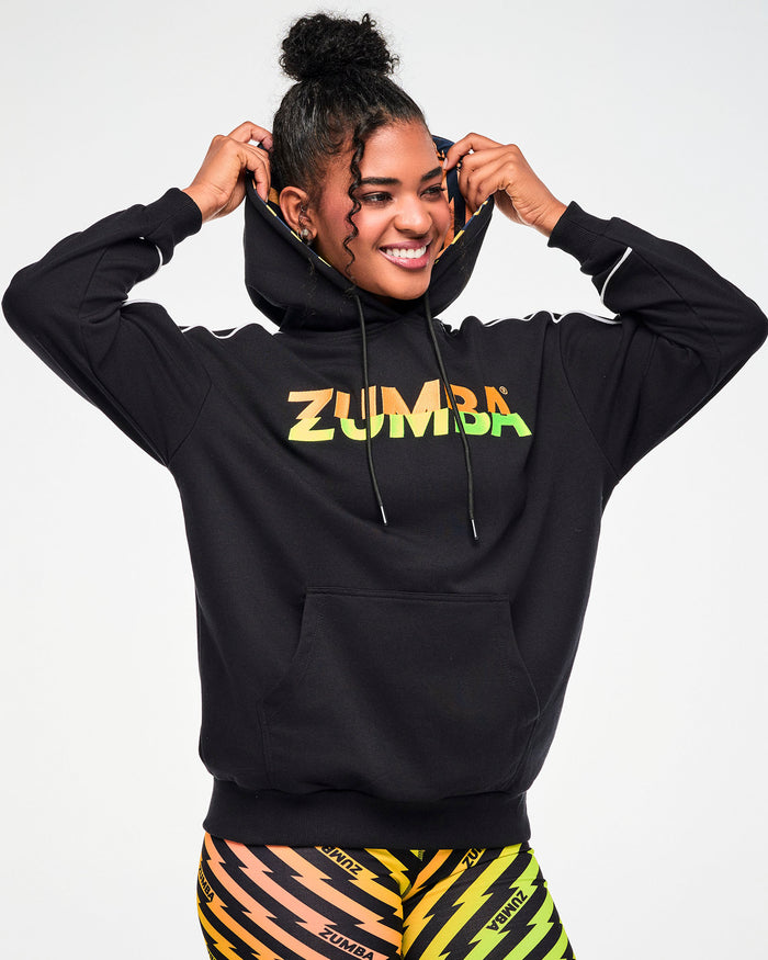 Female model wearing a black ZUMBA hoodie with an orange, yellow, and green ombre lightning bolt-print, paired with matching biker shorts.