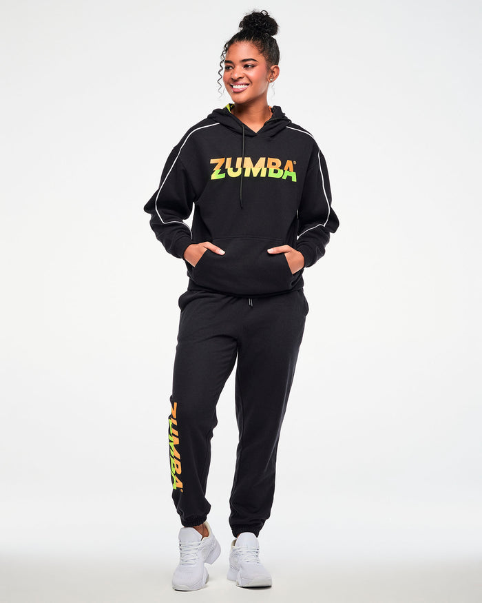 Female model wearing a black hoodie and matching joggers with a gradient "ZUMBA" logo, paired with white sneakers.
