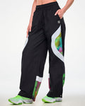 Female model wearing black track pants with a white and multicolor dot-print side panel, hand in her left pocket, showing a black tag featuring a cassette tape icon and "ZUMBA" text, both in white.