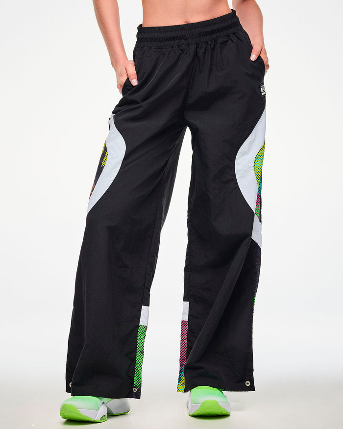 Female model wearing black track pants with a white and multicolor dot-print side panel and green sneakers, hand tucked into the side pocket.
