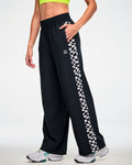 Female model wearing black track pants with a black and white checkerboard side panel and white sneakers, hand in the pocket.