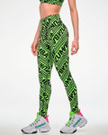 Female model wearing green leggings with a repeated black "ZUMBA" text print and paired with white shoes, side view.