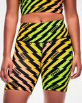 Close-up, side view, of a female model wearing yellow and orange ombre high-waisted biker shorts with a black lightning bolt print and repeated black "ZUMBA" text.