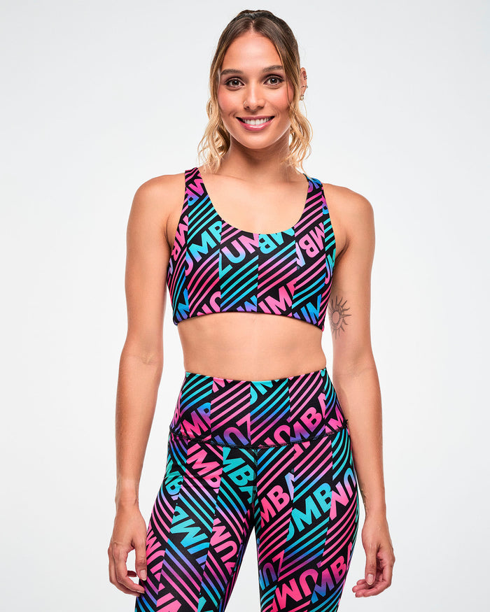 Female model wearing a sports bra with a pink, aqua, and black diagonal "ZUMBA" stripe print and matching leggings.