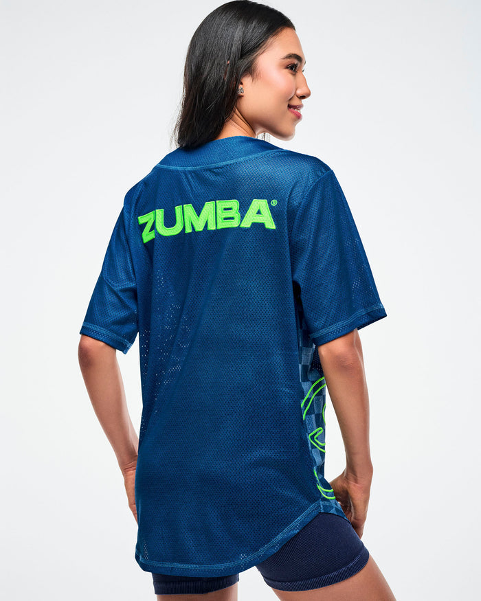 Female model, back view, wearing a blue button-up with “Zumba” in green across the upper back, paired with blue shorts.