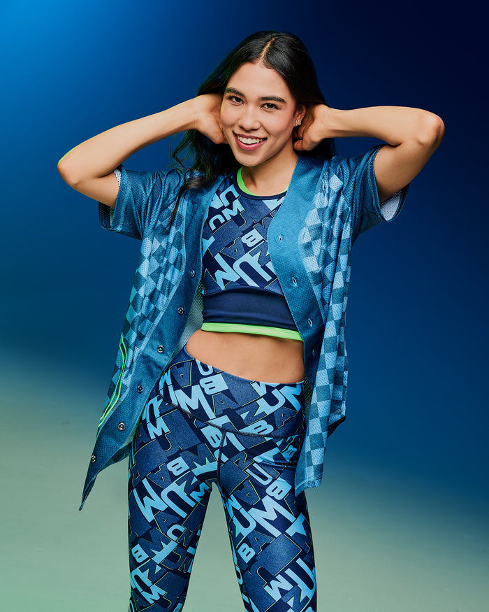 Female model wearing an unbuttoned blue button-up jersey, revealing a patterned bra and matching leggings against a gradient background.