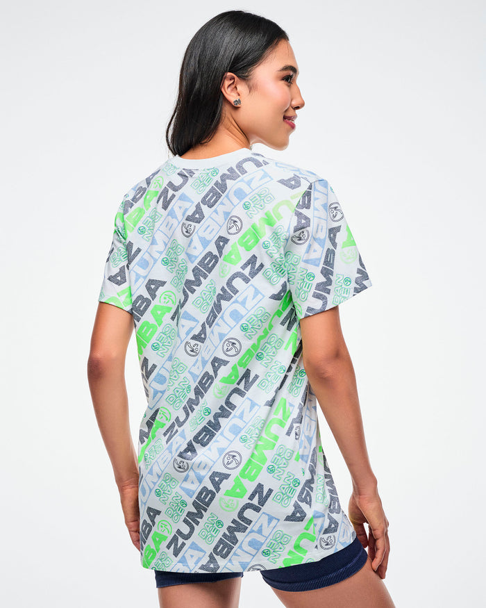 Back view of a female model wearing a Zumba tee with a blue and green 'ZUMBA' logo pattern on a white background.