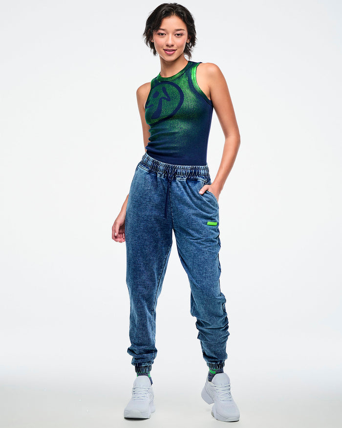 Full view of a female model wearing a green-to-blue gradient tank tucked into blue denim pants and paired with white shoes.