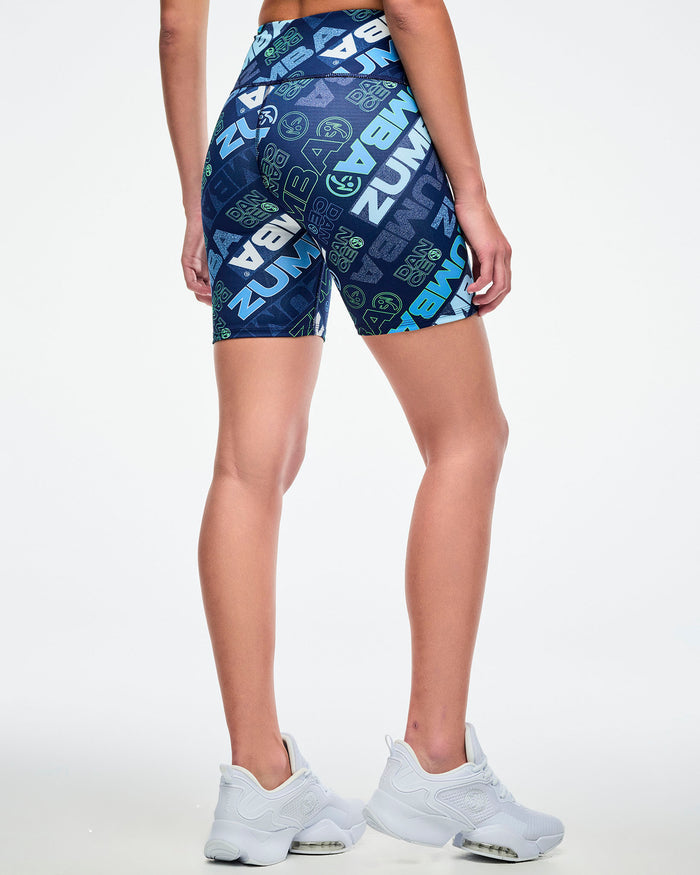 Back view of a female model wearing navy biker shorts with a blue and green “ZUMBA” logo pattern paired with white shoes.