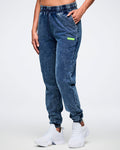 Female model posing with her hand in the left pocket of blue denim pants featuring an elastic waistband and a green “Zumba” label, paired with white shoes.