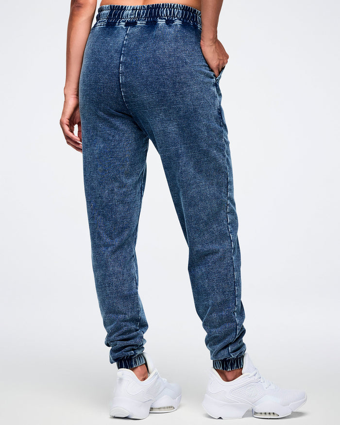 Back view of a female model posing with her hand in the right pocket of blue denim pants featuring an elastic waistband, paired with white shoes.
