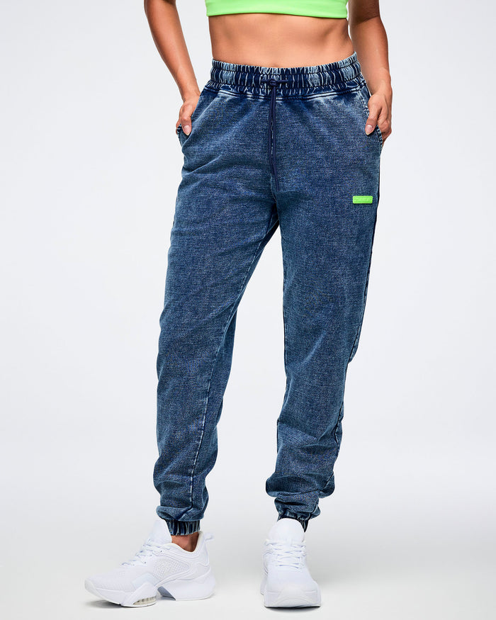 Female model posing with both hands in the pockets of blue denim pants with an elastic waistband and a green “Zumba” label, paired with white shoes.