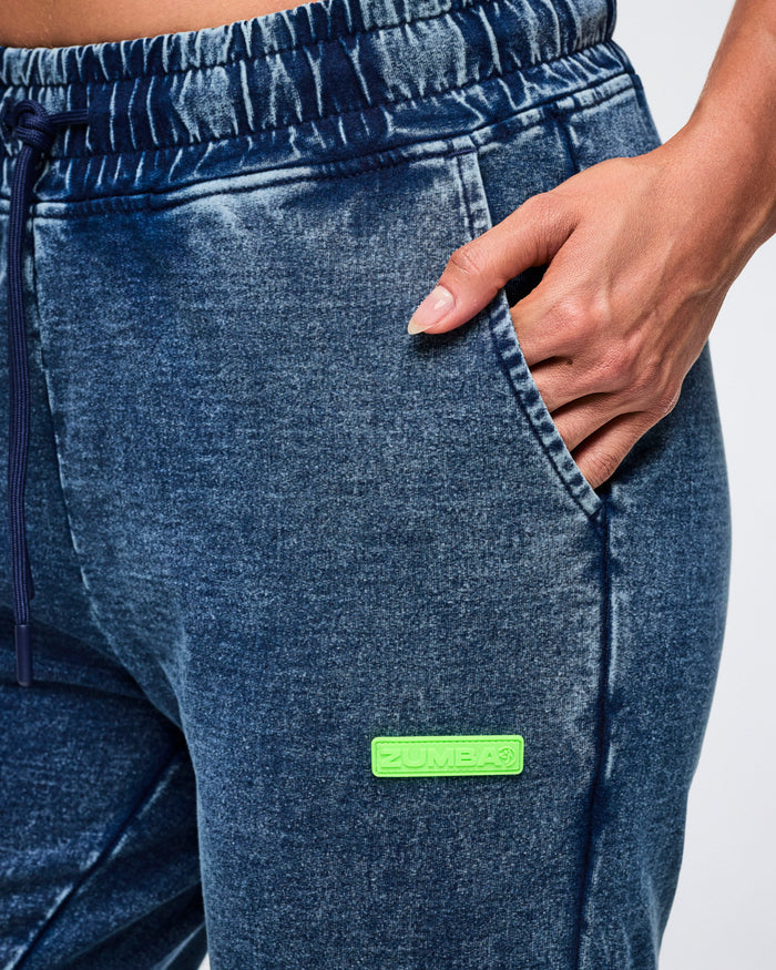 Close-up of a female model with her left hand in the pocket of blue denim pants featuring a green label.