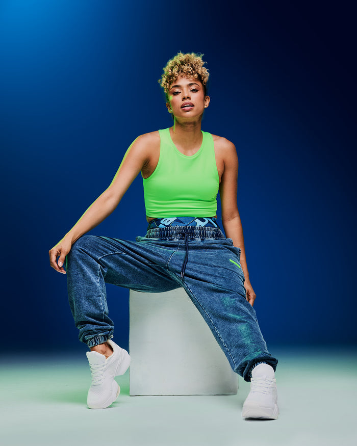 Female model posing on a white box, wearing a neon green crop tank paired with blue denim pants and white shoes on a dark blue background.