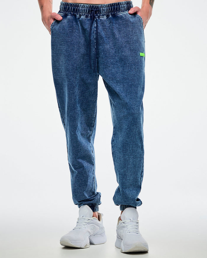 Male model posing with both hands in the pockets of blue pants featuring a green “Zumba” label, paired with white shoes on a white background.