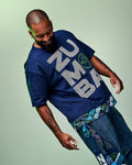 Male model wearing a blue “Zumba” T-shirt with blue denim pants and a bandana with text and graphics hanging from his pocket, set against a light green background.
