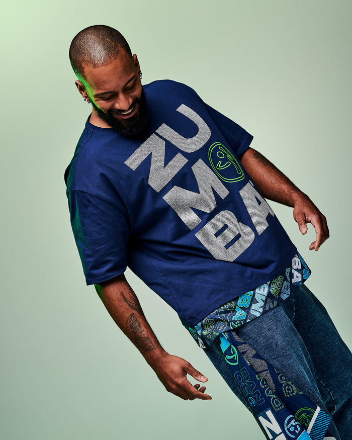 Male model wearing a blue “Zumba” T-shirt with blue denim pants and a bandana with text and graphics hanging from his pocket, set against a light green background.