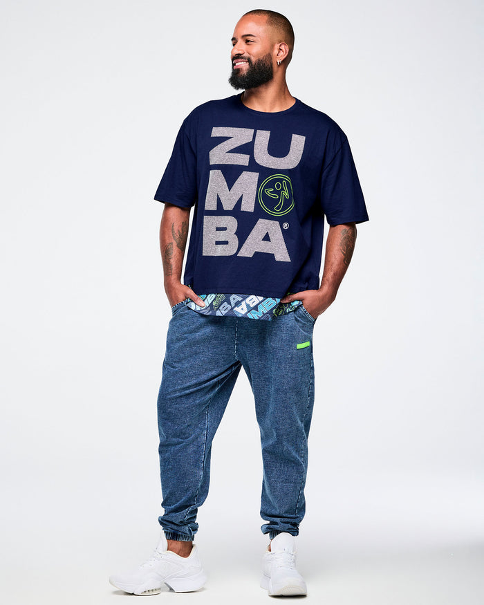 Full view of a male model wearing a blue “Zumba” T-shirt, hands in the pockets of blue denim pants, paired with white shoes.