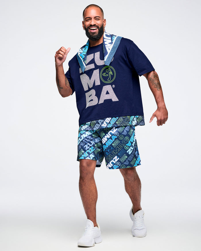 Full view of a male model wearing a blue “Zumba” T-shirt, a bandana draped around his neck, paired with patterned blue “Zumba” shorts and white shoes.