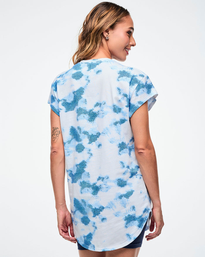 Back view of female model wearing a Zumba Dance Co t-shirt with a blue and white pattern on a plain background.