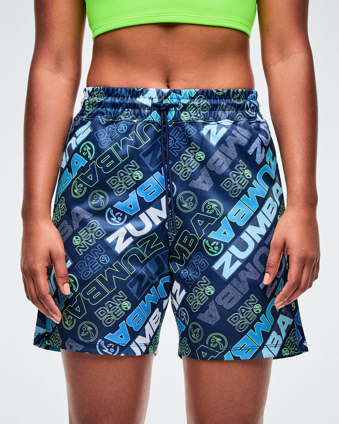 Female model wearing blue patterned Zumba shorts with brand logo on a white background.