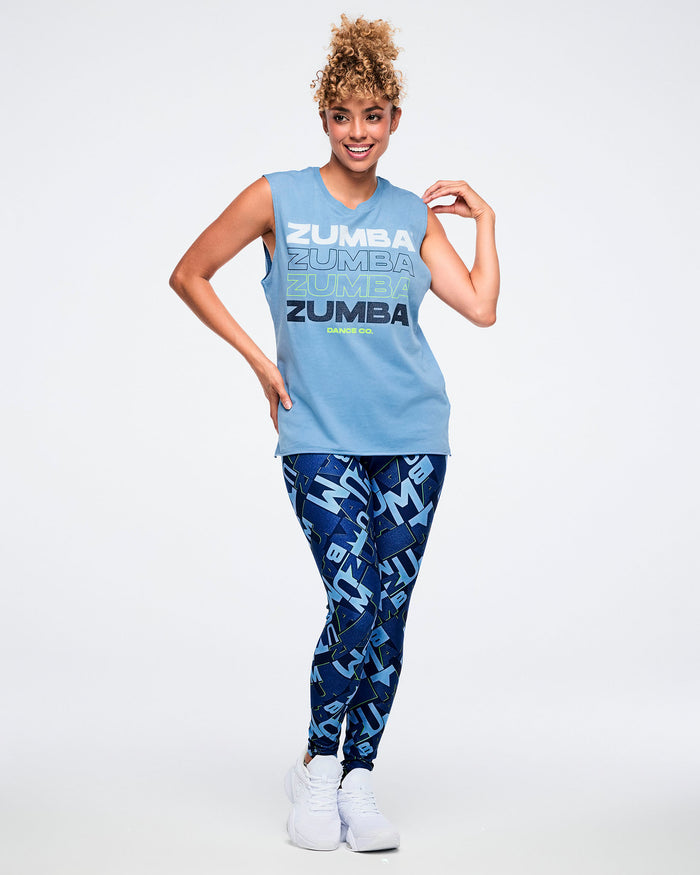 Full body view of a female model wearing a light blue Zumba tank with “ZUMBA” in four text styles and “DANCE CO.” in smaller print paired with blue 'ZUMBA' patterned leggings and white shoes.
