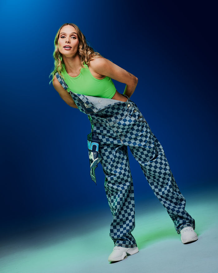 Female model wearing a neon green crop tank and checkered overalls with one strap unfastened, a patterned bandana hanging from her pocket and paired with white shoes against a blue gradient backdrop.