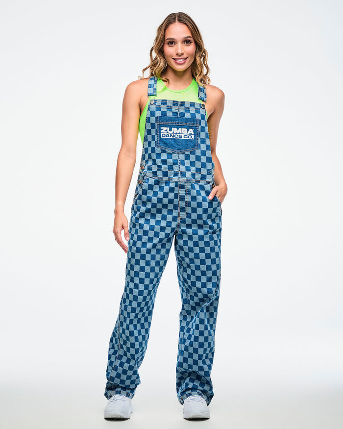Full view of a female model wearing a neon green crop tank under checkered overalls featuring a front pocket with white “Zumba Dance Co.” text, her hand in the left pocket, paired with white shoes.