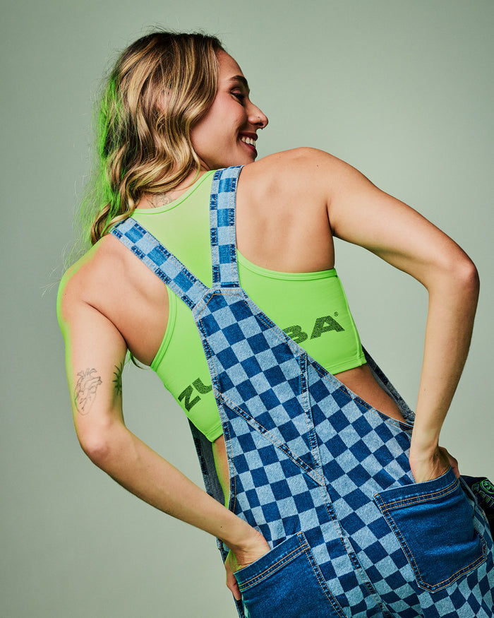 Female model wearing a green crop tank and blue checkered overalls, hands in the plain blue denim back pockets, set against a light green background.